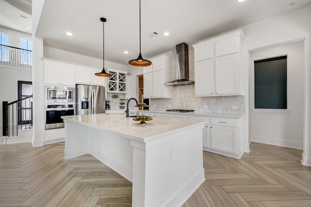 kitchen with an island with sink, light countertops, appliances with stainless steel finishes, wall chimney exhaust hood, and backsplash