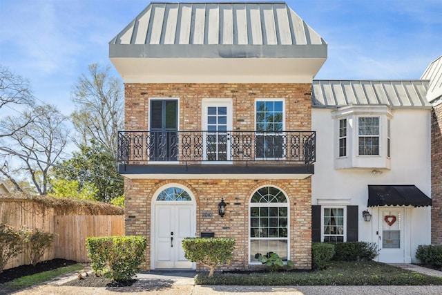 view of front of house with brick siding, stucco siding, a standing seam roof, fence, and a balcony