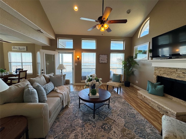 living area featuring a ceiling fan, a stone fireplace, visible vents, wood finished floors, and baseboards