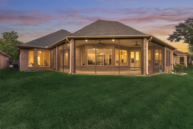 back of house at dusk featuring a sunroom, a yard, a ceiling fan, and brick siding