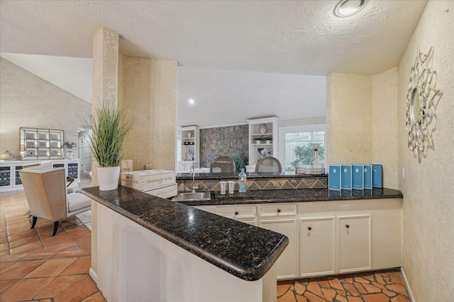 kitchen featuring a textured wall, a peninsula, a sink, vaulted ceiling, and dark stone counters