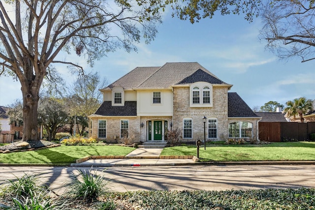 french country home with stucco siding, a shingled roof, a front yard, and fence