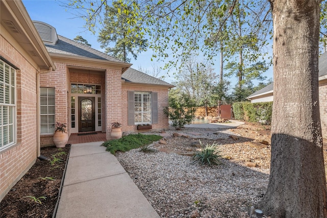 doorway to property with a shingled roof, fence, and brick siding