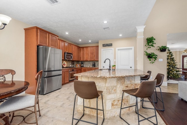 kitchen featuring light stone counters, a sink, appliances with stainless steel finishes, tasteful backsplash, and brown cabinetry