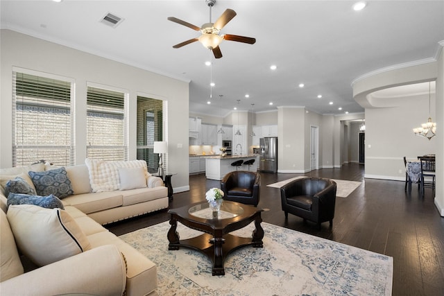 living room featuring ceiling fan with notable chandelier, dark wood-style flooring, visible vents, baseboards, and ornamental molding