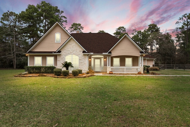 view of front facade featuring stone siding, fence, a yard, covered porch, and a shingled roof