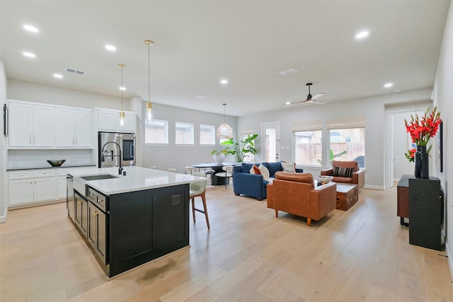 kitchen with a center island with sink, visible vents, white cabinetry, a sink, and light wood-type flooring