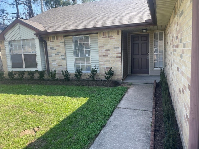 view of exterior entry with a shingled roof, a lawn, and brick siding