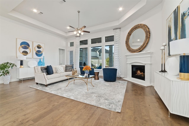 living area with a tray ceiling, recessed lighting, a glass covered fireplace, ceiling fan, and wood finished floors