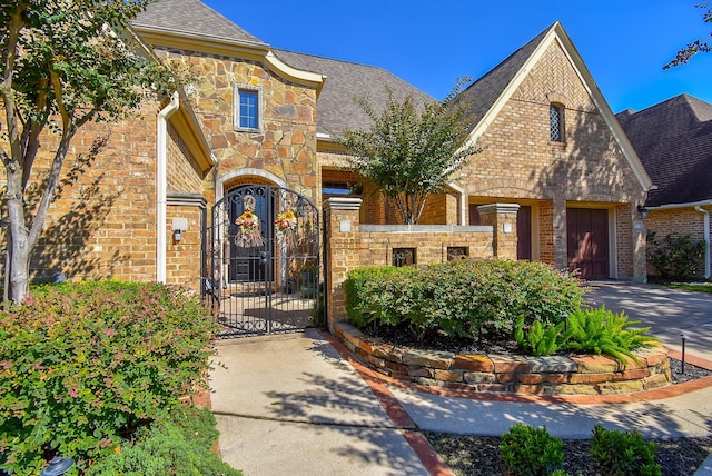 tudor home featuring a garage, brick siding, stone siding, driveway, and a gate