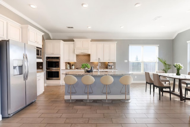 kitchen with stainless steel appliances, a sink, ornamental molding, decorative backsplash, and an island with sink