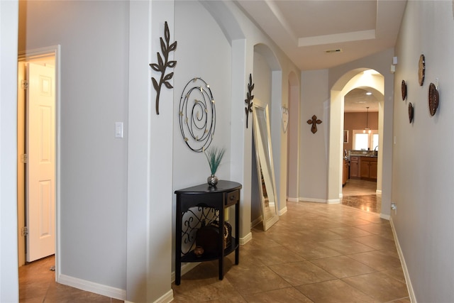 hallway featuring arched walkways, light tile patterned floors, visible vents, baseboards, and a tray ceiling