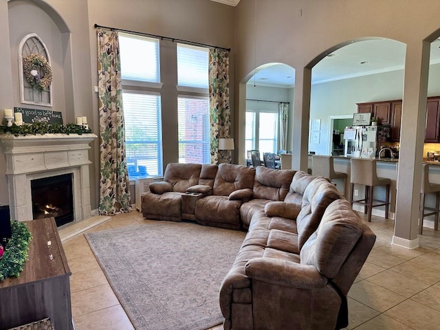 living area featuring arched walkways, a towering ceiling, a premium fireplace, ornamental molding, and light tile patterned flooring