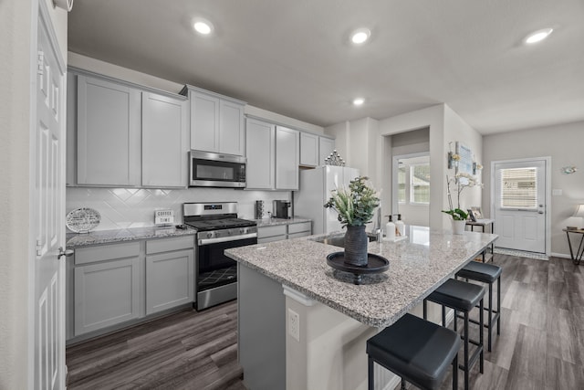 kitchen with gray cabinetry, dark wood-type flooring, appliances with stainless steel finishes, decorative backsplash, and an island with sink