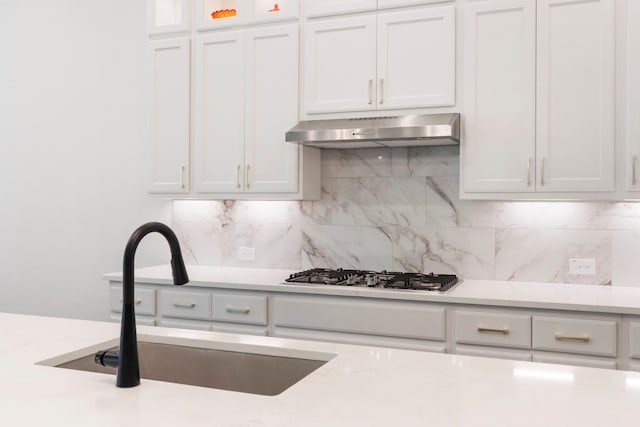 kitchen featuring tasteful backsplash, white cabinets, under cabinet range hood, stainless steel gas cooktop, and a sink