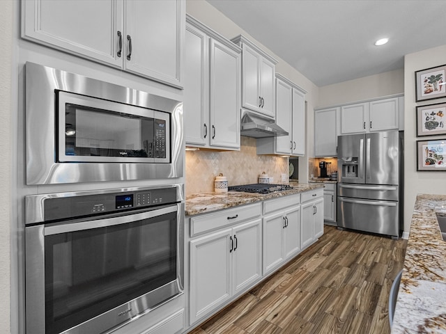 kitchen with light stone counters, under cabinet range hood, stainless steel appliances, tasteful backsplash, and dark wood finished floors