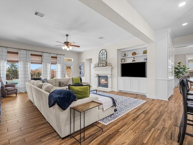 living area with visible vents, a glass covered fireplace, ceiling fan, wood finished floors, and built in shelves