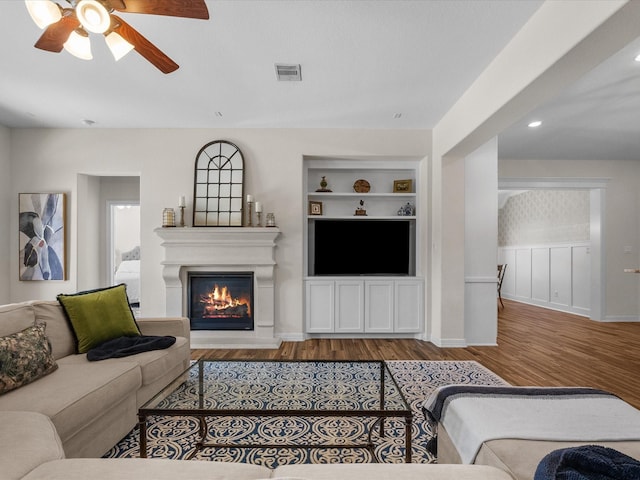 living room with visible vents, a ceiling fan, a glass covered fireplace, wood finished floors, and built in shelves