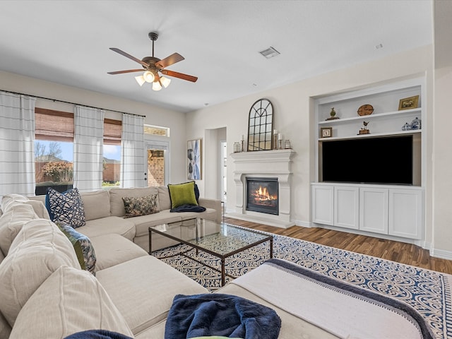 living area featuring built in shelves, wood finished floors, visible vents, baseboards, and a glass covered fireplace