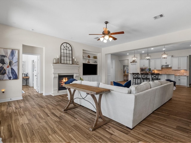 living room with dark wood-style floors, built in shelves, visible vents, a glass covered fireplace, and baseboards