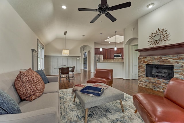 living room featuring arched walkways, a ceiling fan, lofted ceiling, dark wood-style floors, and a stone fireplace