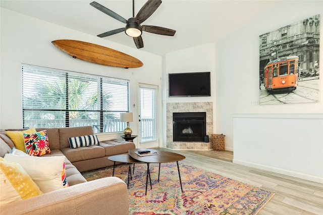 living area with ceiling fan, a tiled fireplace, wood finished floors, and baseboards