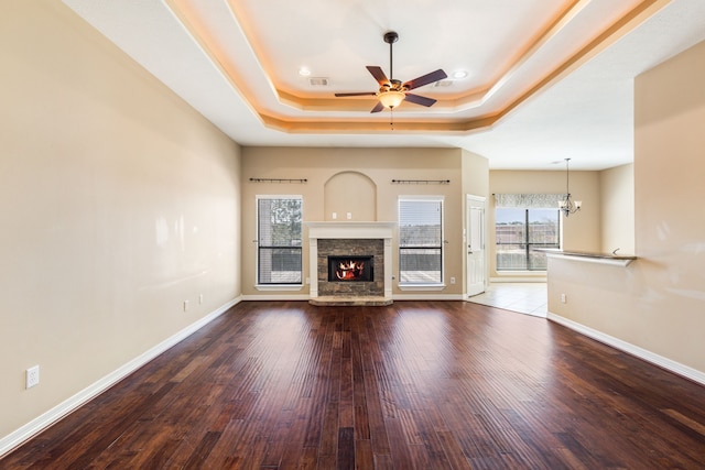 unfurnished living room with a raised ceiling, ceiling fan with notable chandelier, wood finished floors, a stone fireplace, and baseboards