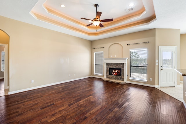 unfurnished living room with ceiling fan, a stone fireplace, wood finished floors, arched walkways, and a raised ceiling