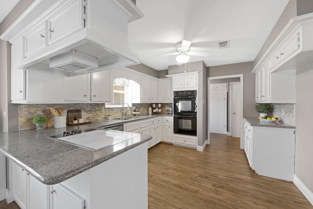 kitchen with custom exhaust hood, dobule oven black, white cabinetry, a sink, and a peninsula