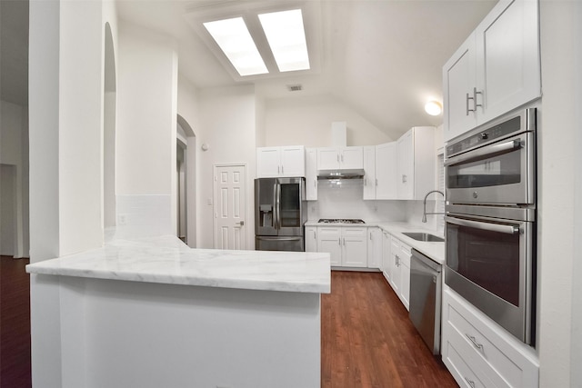 kitchen with vaulted ceiling with skylight, light stone counters, stainless steel appliances, a peninsula, and a sink