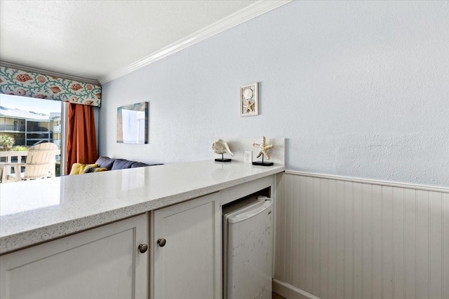 kitchen with a wainscoted wall, crown molding, a textured wall, white cabinets, and light stone countertops