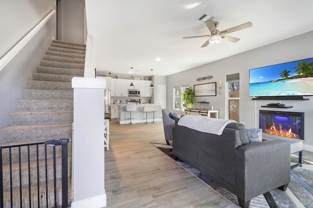 living area with visible vents, a ceiling fan, a glass covered fireplace, light wood-style flooring, and stairway