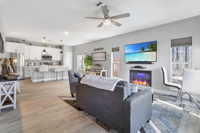 living room with light wood finished floors, baseboards, visible vents, a ceiling fan, and a glass covered fireplace