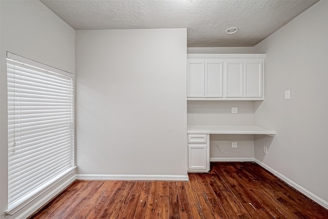 unfurnished office with a textured ceiling, baseboards, built in desk, and wood finished floors