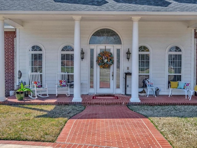 view of exterior entry with a porch, a lawn, a shingled roof, and brick siding