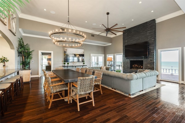 dining space featuring ceiling fan, a fireplace, a towering ceiling, ornamental molding, and dark wood finished floors