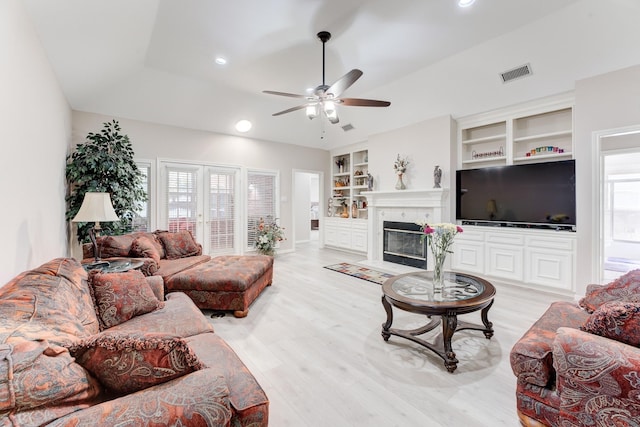 living area featuring built in features, visible vents, light wood-style flooring, a ceiling fan, and a glass covered fireplace