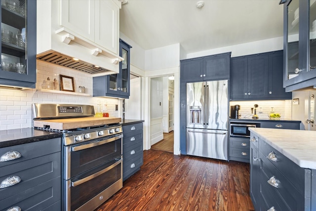 kitchen featuring light stone counters, stainless steel appliances, tasteful backsplash, dark wood-type flooring, and glass insert cabinets