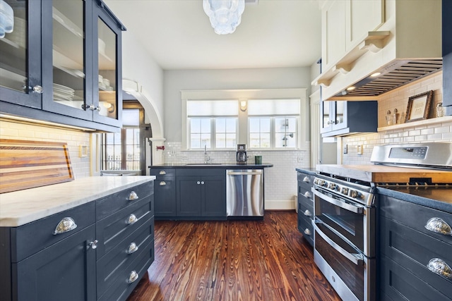 kitchen with dark wood finished floors, stainless steel appliances, glass insert cabinets, a sink, and under cabinet range hood