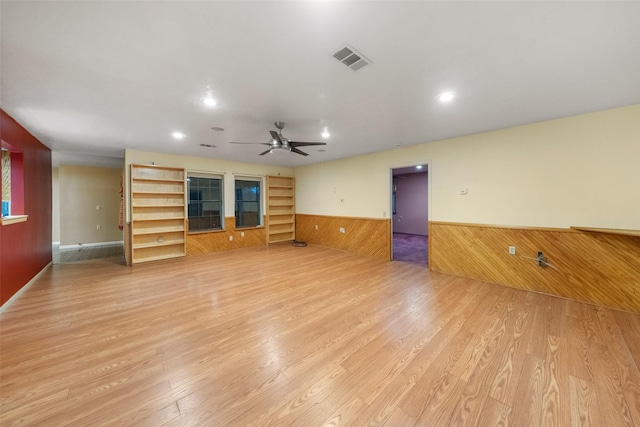 unfurnished living room featuring wainscoting, light wood-type flooring, visible vents, a ceiling fan, and wood walls