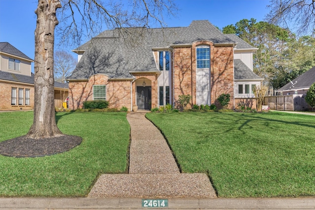 french country inspired facade featuring a front lawn, roof with shingles, fence, and brick siding