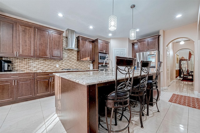 kitchen featuring arched walkways, stainless steel microwave, wall chimney range hood, and light tile patterned floors