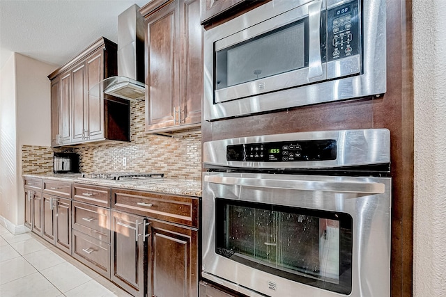 kitchen featuring light tile patterned floors, decorative backsplash, wall chimney exhaust hood, appliances with stainless steel finishes, and light stone countertops