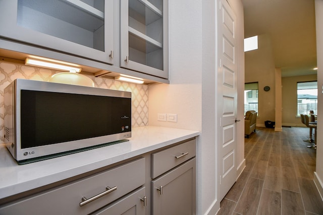 kitchen featuring gray cabinetry, dark wood-type flooring, decorative backsplash, stainless steel microwave, and glass insert cabinets