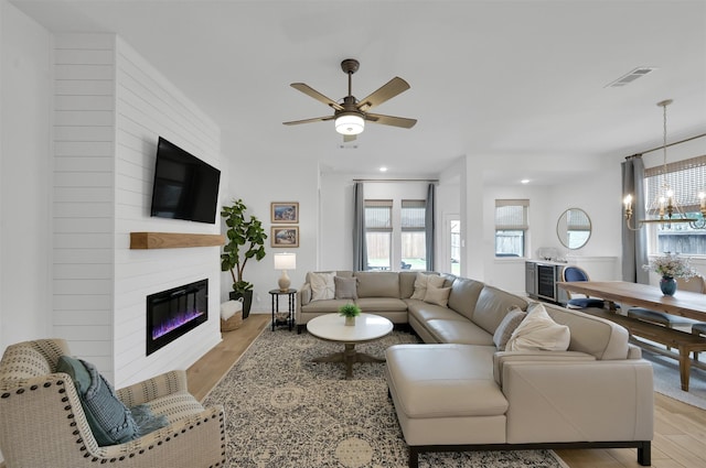 living room featuring a large fireplace, visible vents, light wood-style flooring, ceiling fan with notable chandelier, and recessed lighting