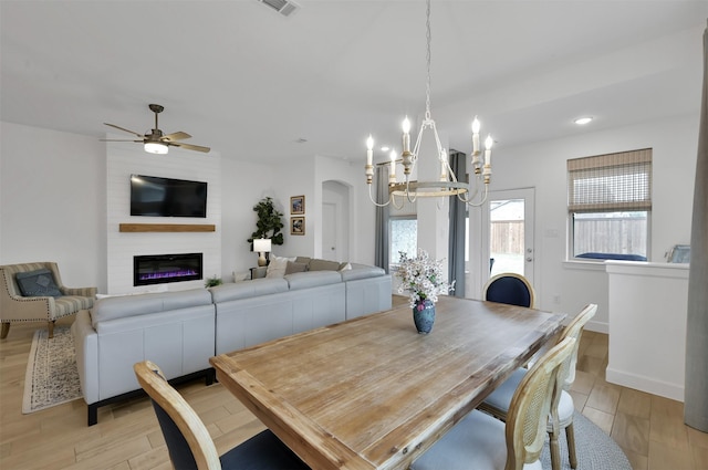 dining room with light wood finished floors, baseboards, a large fireplace, and ceiling fan with notable chandelier