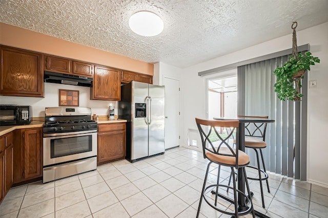 kitchen featuring light countertops, appliances with stainless steel finishes, brown cabinetry, light tile patterned flooring, and under cabinet range hood