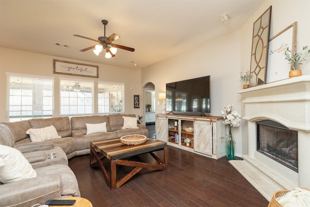 living area featuring arched walkways, a fireplace with raised hearth, wood finished floors, visible vents, and a ceiling fan