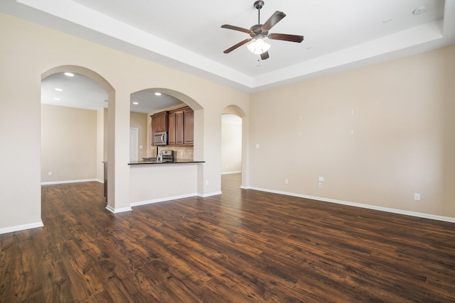 unfurnished living room featuring dark wood-style floors, arched walkways, a raised ceiling, ceiling fan, and baseboards