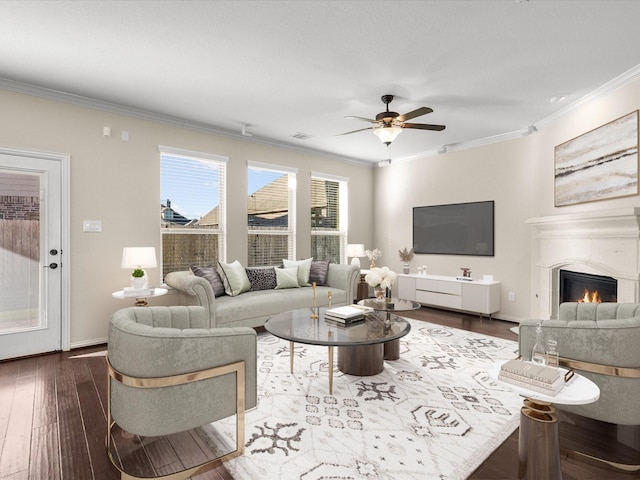 living room with dark wood-style flooring, ornamental molding, a glass covered fireplace, a ceiling fan, and baseboards